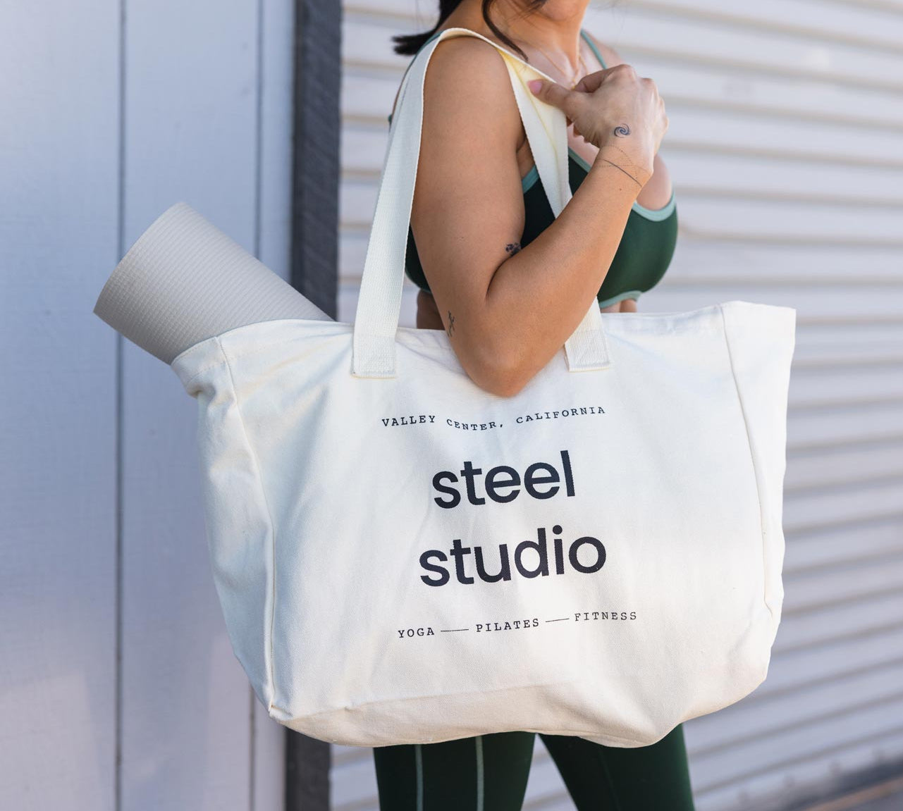 A woman in green activewear holds a large white canvas tote bag printed with 'steel studio' in black text, standing in front of a white corrugated metal wall.
